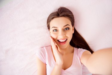 Self top above view portrait of pretty, brunette, charming, cute, glad, pleased, positive girl holding palm near cheek looking at camera, shooting selfie on front camera, lying on back on bed