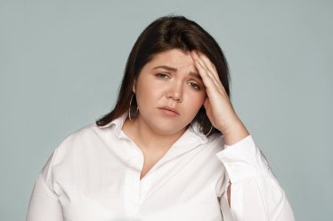 Studio shot of upset frustrated young chubby female employee wearing formal shirt holding hand on her forehead, having mournful look, suffering from headache or hangover after sleepless night