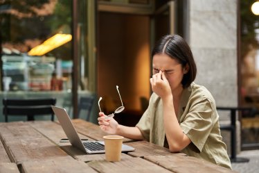 Young asian woman feeling tired after working with laptop, sitting in cafe on bench outdoors, drinking coffee, looking exhausted.