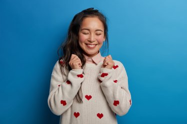 Optimistic Asian girl raises clenched fists, anticipates for good results of exam, closes eyes and smiles broadly, has pony tail, wears sweater isolated on blue background. People, emotions, lifestyle