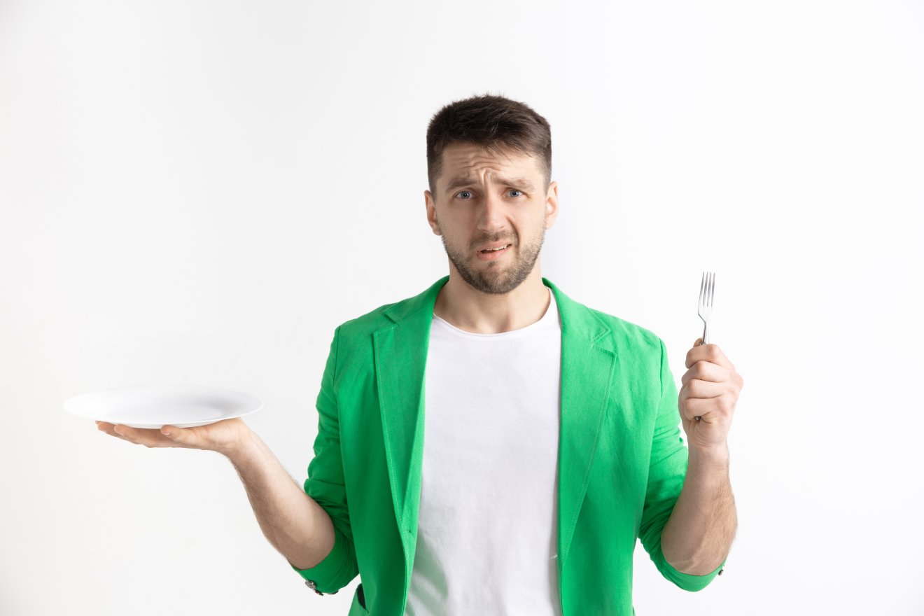 Young sad attractive caucasian guy holding empty dish and fork isolated on grey background. Copy space and mock up. Blank template background. Reject, rejection concept