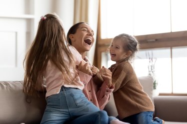 Cheerful positive family cute small funny kids children daughters enjoying tickling playing lifestyle game with young happy mum embracing laughing having fun relaxing sitting on sofa at home together