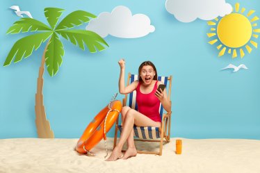 Overemotive young woman holds modern cell phone, clenches fist with triumph, rejoices getting good news on email, rests outdoor at beach, sits at deck chair, lifebuoy and sunscreen lotion near
