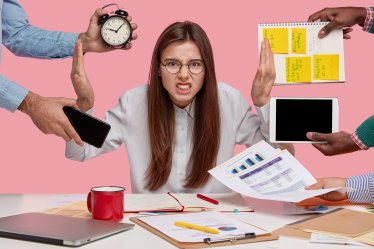 Overworked young employee refuses all things, frowns face in annoyance, sits at desktop with paper documents and notepad, isolated over pink background. Female workes bothered by many questions